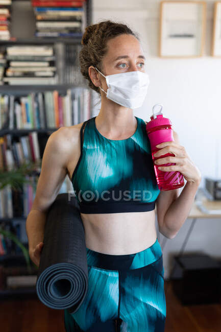 Белая женщина проводит время дома в спортивной одежде и маске против коронавируса, ковид 19, держит мат и пластиковую бутылку. Социальное дистанцирование и самоизоляция в карантинной изоляции. — стоковое фото