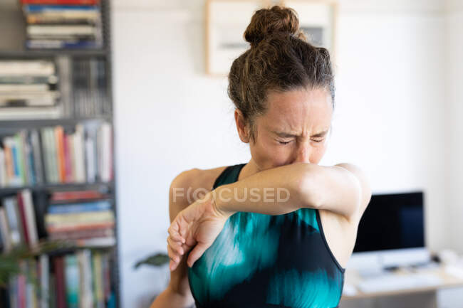 Белая женщина проводит время дома в спортивной одежде, прикрывая лицо во время кашля. Социальное дистанцирование и самоизоляция в карантинной изоляции. — стоковое фото