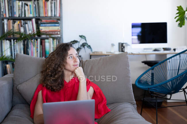 Кавказька жінка проводить час удома, одягнувши рожеву сукню, сидячи на дивані і використовуючи свій портативний комп'ютер. Соціальна дистанція і самоізоляція в карантинному блокуванні.. — стокове фото