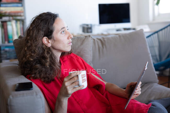 Femme caucasienne passant du temps à la maison, portant une robe rose, assise sur un canapé, tenant une tasse de café et utilisant sa tablette. Distance sociale et isolement personnel en quarantaine. — Photo de stock