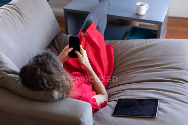 Femme caucasienne passant du temps à la maison, portant une robe rose, assise sur un canapé et utilisant son smartphone. Distance sociale et isolement personnel en quarantaine. — Photo de stock
