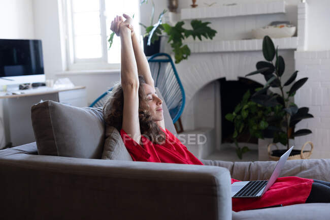 Кавказька жінка проводить час удома, одягнувши рожеву сукню, сидячи на дивані і користуючись ноутбуком, роблячи перерву і розтягуючи руки. Соціальна дистанція і самоізоляція в карантинному блокуванні.. — стокове фото
