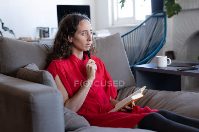 Femme caucasienne passant du temps à la maison, portant une robe rose, assise sur un canapé et lisant un livre. Distance sociale et isolement personnel en quarantaine. — Photo de stock