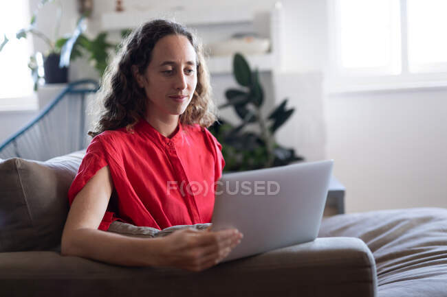 Femme caucasienne passant du temps à la maison, portant une robe rose, assise sur un canapé et utilisant son ordinateur portable. Distance sociale et isolement personnel en quarantaine. — Photo de stock