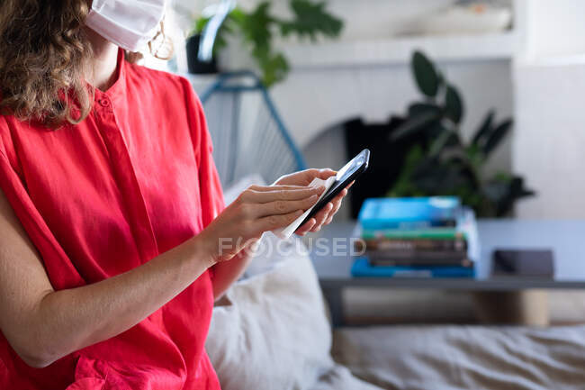Средняя часть белой женщины проводит время дома, одетая в розовое платье и маску для лица против коронавируса, ковид 19, чистит смартфон. Социальное дистанцирование и самоизоляция в карантинной изоляции. — стоковое фото