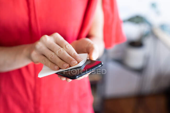 Mid section of woman spending time at home, wearing a pink dress, cleaning her smartphone. Social distancing and self isolation in quarantine lockdown. — Stock Photo
