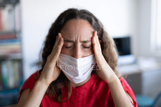 Кавказька жінка проводить час удома, одягнувши рожеву сукню і маску обличчя проти коронавірусу, ковидка 19, страждаючи від головного болю. Соціальна дистанція і самоізоляція в карантинному блокуванні.. — стокове фото