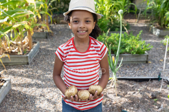 Niña afroamericana distanciamiento social en casa durante el cierre de cuarentena, de pie en un jardín, sonriendo y presentando patatas frescas. - foto de stock
