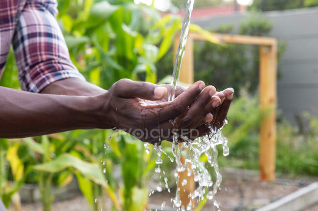 Partie médiane d'un homme afro-américain prenant ses distances sociales à la maison pendant le confinement en quarantaine, se lavant les mains avec un robinet de jardin. — Photo de stock