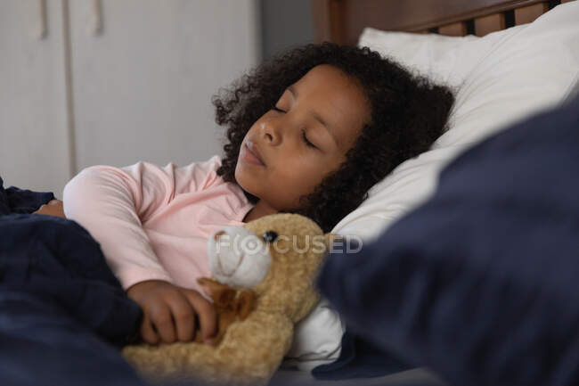 Афроамериканська дівчинка спить у спальні свого батька і обіймає плюшевого ведмедика, під час соціального дистанціювання вдома під час карантину.. — стокове фото