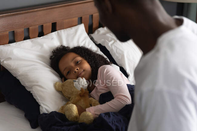 Homem afro-americano acordando sua filha, dormindo em sua cama e abraçando um ursinho de pelúcia, durante o distanciamento social em casa durante o bloqueio de quarentena . — Fotografia de Stock