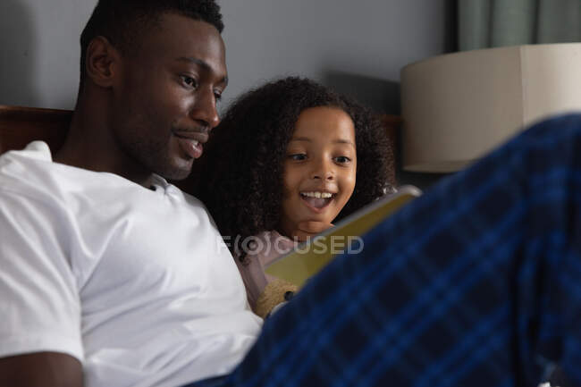 Afro-américaine fille et son père social distanciation à la maison pendant la quarantaine verrouillage, passer du temps ensemble, avoir du plaisir et lire un livre dans un lit. — Photo de stock