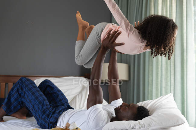 Afro-americano menina e seu pai social distanciamento em casa durante quarentena bloqueio, passar tempo juntos, se divertindo e jogar em uma cama . — Fotografia de Stock