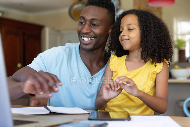 Menina afro-americana vestindo uma blusa amarela, distanciamento social em casa durante o bloqueio de quarentena, passando tempo com seu pai usando um computador portátil . — Fotografia de Stock