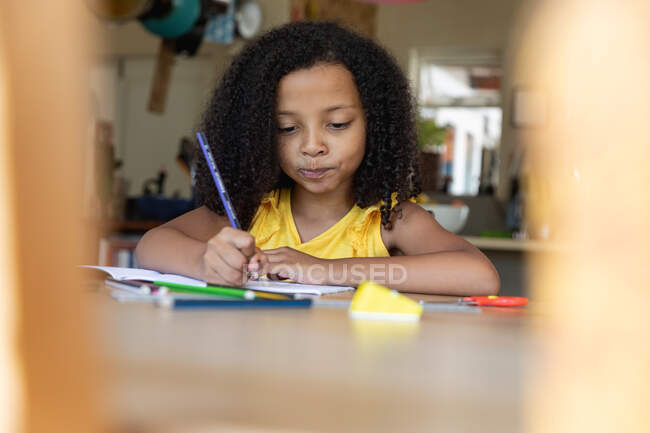 Menina afro-americana vestindo uma blusa amarela, distanciamento social em casa durante o confinamento de quarentena, sentado a uma mesa e desenhando imagens . — Fotografia de Stock