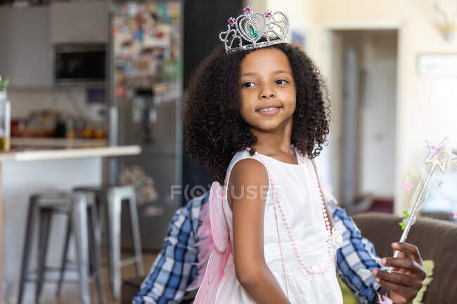 Ragazza afroamericana che indossa una corona giocattolo, distanza sociale a casa durante il blocco di quarantena, giocare con il padre in un salotto. — Foto stock