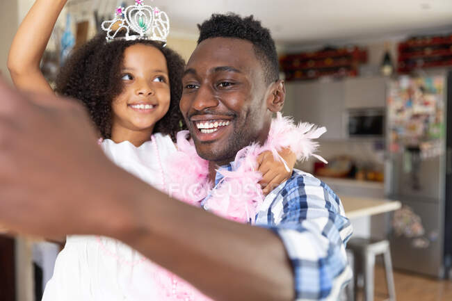 Menina afro-americana usando asas de fada de brinquedo, distanciamento social em casa durante o bloqueio de quarentena, brincando com seu pai vestindo uma boa rosa e tirando selfies . — Fotografia de Stock