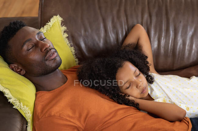 Menina afro-americana e seu pai, distanciamento social em casa durante o confinamento de quarentena, tirando uma soneca em um sofá de couro . — Fotografia de Stock