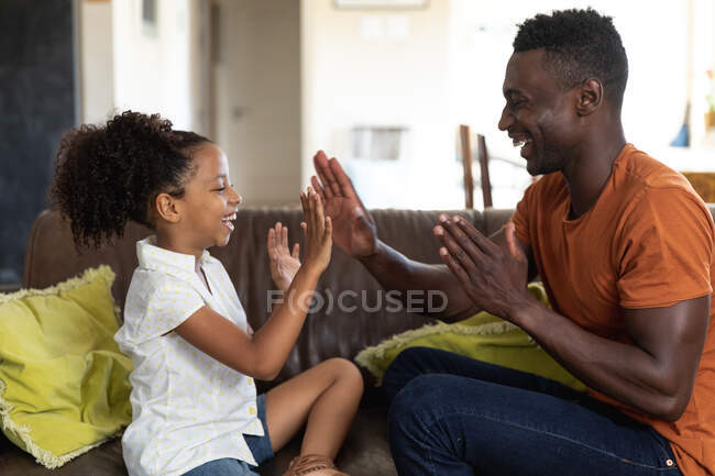 African American girl and her father, social distancing at home during quarantine lockdown, sitting on a couch and playing together, clapping their hands. — Stock Photo