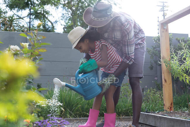 Niña afroamericana y su padre social distanciándose en casa durante el cierre de cuarentena, pasando tiempo en su jardín juntos, regando flores, en un día soleado. - foto de stock