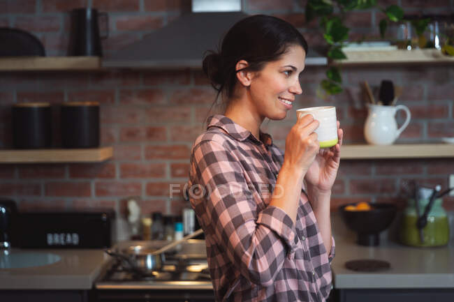 Frau mit gemischter Rasse, die während der Coronavirus-Epidemie 19 Zeit zu Hause mit Selbstisolierung und sozialer Distanzierung in Quarantäne verbringt und in der Küche eine Tasse Kaffee hält. — Stockfoto