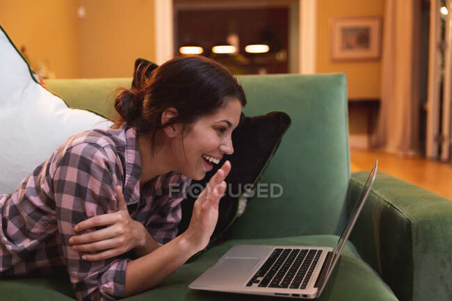 Mujer de raza mixta pasar tiempo en casa auto aislamiento y distanciamiento social en cuarentena de bloqueo durante coronavirus covid 19 epidemia, acostado en un sofá con el ordenador portátil ondeando sonriendo en la sala de estar. - foto de stock