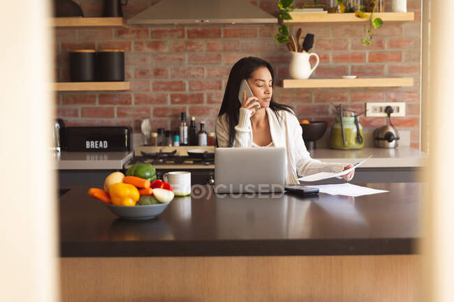 Mixed race woman spending time at home, working from home using laptop computer in kitchen. Self isolating and social distancing in quarantine lockdown during coronavirus covid 19 epidemic. — Stock Photo