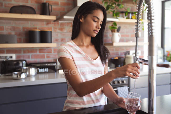 Смешанная расовая женщина проводит время дома самоизолируясь и социальное дистанцирование в карантинной изоляции во время эпидемии коронавируса ковида 19, разливая воду в стекло на кухне. — стоковое фото
