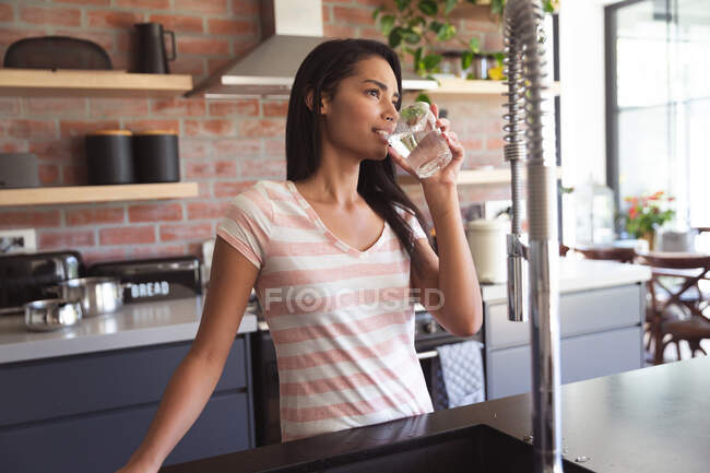 Mixed race woman spending time at home self isolating and social distancing in quarantine lockdown during coronavirus covid 19 epidemic, drinking water in kitchen. — Stock Photo