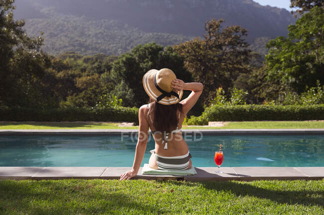 Вид ззаду змішаної раси жінка проводить час за допомогою самоізоляції басейну та соціального дистанціювання в карантинному замку під час епідемії коронавірусу, сидячи біля басейну в саду . — стокове фото