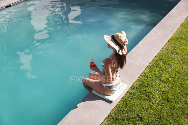 Смешанная расовая женщина, проводящая время у бассейна самоизоляция и социальное дистанцирование в карантинной изоляции во время эпидемии коронавируса, сидя у бассейна, держа напиток. — стоковое фото
