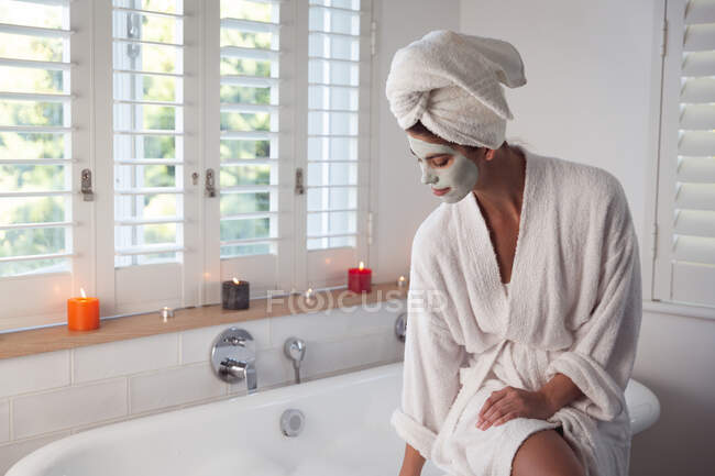 Змішана расова жінка проводить час удома, сидячи на ванні з маскою для бігу в ванній кімнаті. Самоізоляція і соціальна дистанція в карантинній блокаді під час коронавірусу covid 19 епідемії. — стокове фото