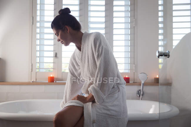 Змішана жінка проводить час вдома, сидячи на ванні з маскою для обличчя під час ванни у ванній кімнаті. Самоізоляція та соціальне дистанціювання в карантинному блокуванні під час епідемії коронавірусу 19 . — стокове фото