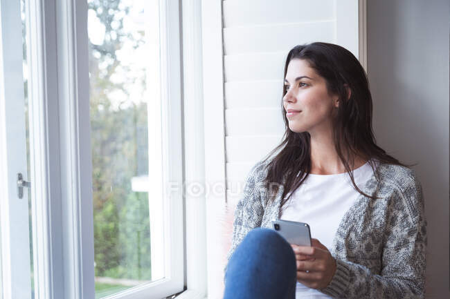Смешанная расовая женщина проводит время дома, сидя на оконном сидении, держа смартфон в гостиной. Самоизоляция и социальное дистанцирование в карантинной изоляции во время эпидемии коронавируса ковида 19. — стоковое фото
