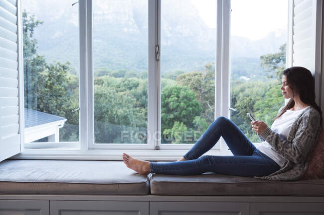 Змішана жінка проводить час вдома, сидячи на віконному сидінні, тримаючи смартфон у вітальні. Самоізоляція та соціальне дистанціювання в карантинному блокуванні під час епідемії коронавірусу 19 . — стокове фото