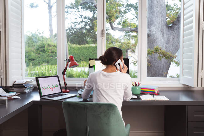 Rear view of mixed race woman spending time at home self isolating and social distancing in quarantine lockdown during coronavirus covid 19 epidemic, sitting at desk using smartphone working from home — Stock Photo