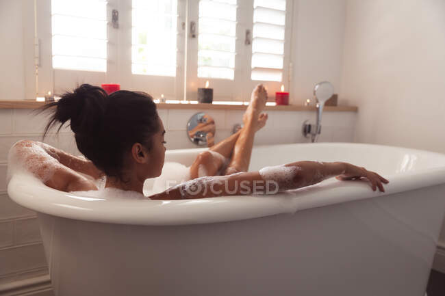 Mixed race woman spending time at home self isolating and social distancing in quarantine lockdown during coronavirus covid 19 epidemic, lying in bathtub relaxing in bathroom. — Stock Photo