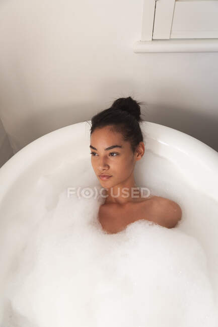Gemischte Rassenfrau, die während der Coronavirus-Epidemie 19 in der Badewanne liegt, die mit Schaumstoff bedeckt ist und sich im Badezimmer entspannt, verbringt Zeit zu Hause mit Selbstisolation und sozialer Distanzierung. — Stockfoto