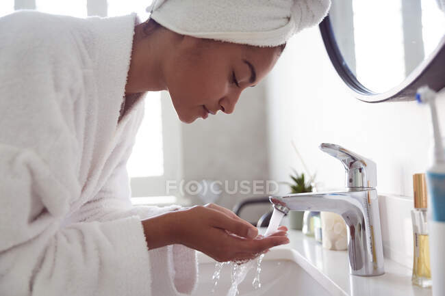 Смешанная расовая женщина проводит время дома самоизоляция и социальное дистанцирование в карантинной изоляции во время эпидемии коронавируса ковид 19, одетая в халат с полотенцем на голове, моющая в ванной комнате. — стоковое фото