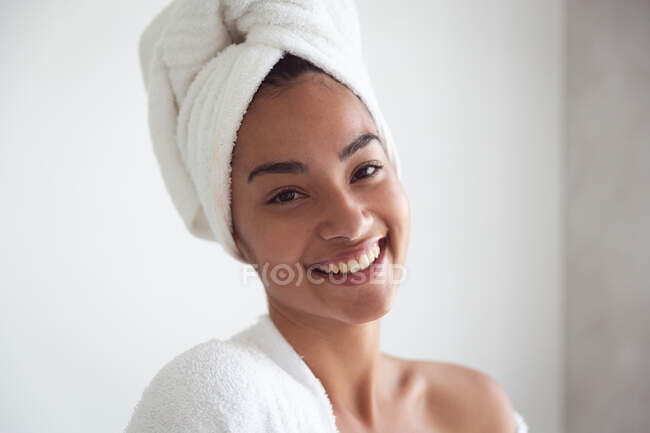 Portrait of mixed race woman spending time at home self isolating and social distancing in quarantine lockdown during coronavirus covid 19 epidemic, wearing bathrobe with towel on her head in bathroom — Stock Photo