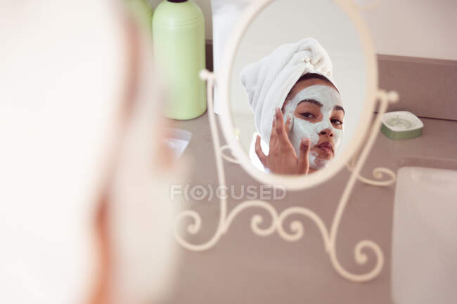 Mixed race woman spending time at home self isolating and social distancing in quarantine lockdown during coronavirus covid 19 epidemic, looking at mirror with face mask on in bathroom. — Stock Photo