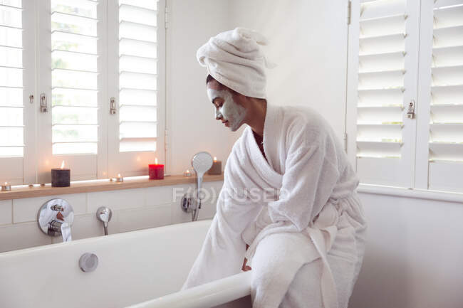 Mixed race woman spending time at home self isolating and social distancing in quarantine lockdown during coronavirus covid 19 epidemic, sitting on bathtub with face mask on running bath in bathroom. — Stock Photo