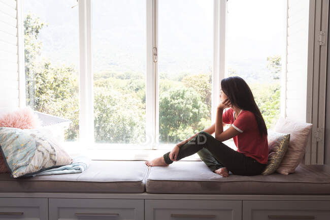 Gemischte Rassenfrau, die während der Coronavirus-Epidemie des 19. Jahrhunderts Zeit zu Hause mit Selbstisolierung und sozialer Distanzierung in Quarantäne verbringt, sitzt auf Fensterbank und schaut im Wohnzimmer aus dem Fenster. — Stockfoto