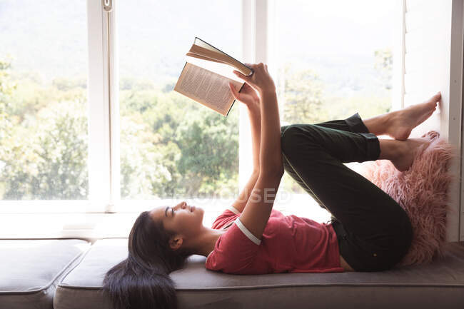 Mixed race woman spending time at home self isolating and social distancing in quarantine lockdown during coronavirus covid 19 epidemic, lying on window seat reading a book in sitting room. — Stock Photo