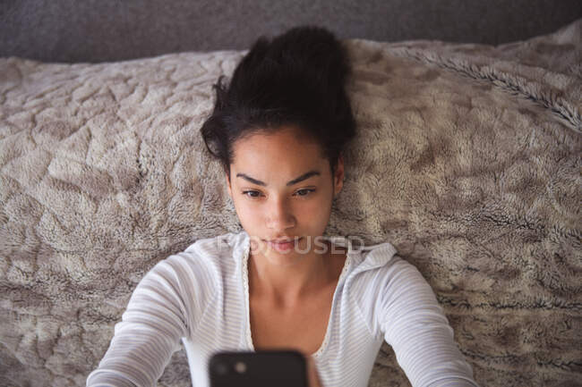 Gemischte Rassenfrau, die während der Coronavirus-Epidemie 19 in Quarantäne lebt und sich zu Hause selbst isoliert und sozial distanziert, liegt im Bett und macht Selfies mit ihrem Smartphone im Schlafzimmer. — Stockfoto