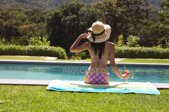 Rear view of mixed race woman spending time by pool self isolating and social distancing in quarantine lockdown during coronavirus covid 19 epidemic, sitting by a swimming pool holding a drink. — Stock Photo