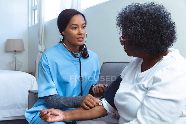Senior Mixed Race Frau verbringt Zeit zu Hause, wird von einer Mixed Race Krankenschwester besucht, die Krankenschwester nimmt Blutdruck — Stockfoto