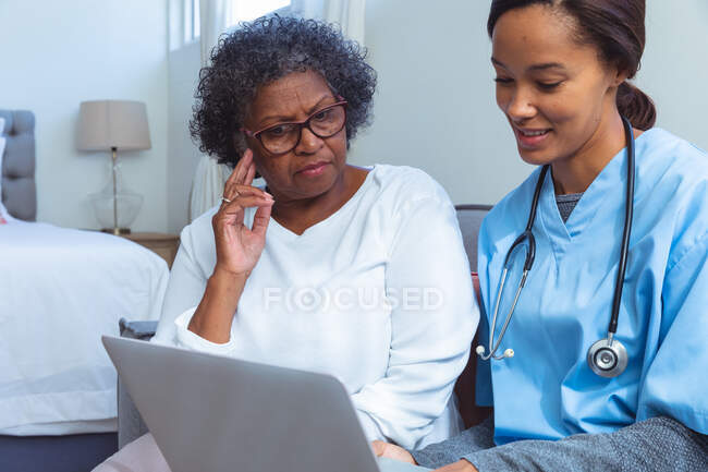 Femme âgée métisse qui passe du temps à la maison, visitée par une infirmière métisse, l'infirmière utilisant un ordinateur portable — Photo de stock