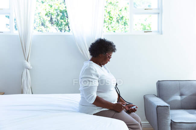 Senior mixed race woman spending time at home, social distancing and self isolation in quarantine lockdown, sitting on a bed and taking her blood pressure — Stock Photo