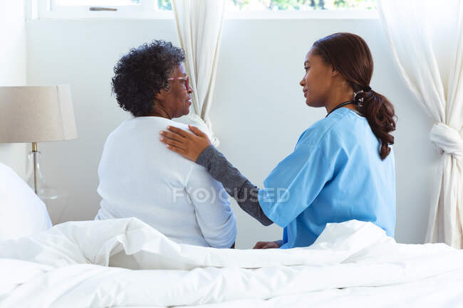 Seniorin mit gemischter Rasse verbringt Zeit zu Hause, wird von einer Krankenschwester mit gemischter Rasse besucht, sitzt auf einem Bett und redet — Stockfoto